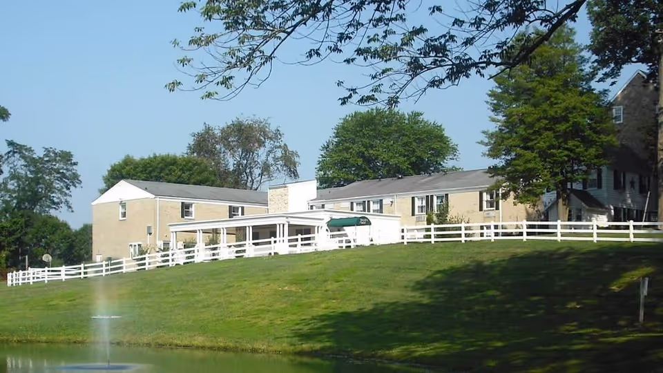Exterior view of Jacob's Well Assisted Living facility showing a two-story building with beige brick walls and white trim, surrounded by green grass, trees, and a white fence. A small pond with a water fountain is visible in the foreground under a clear blue sky.