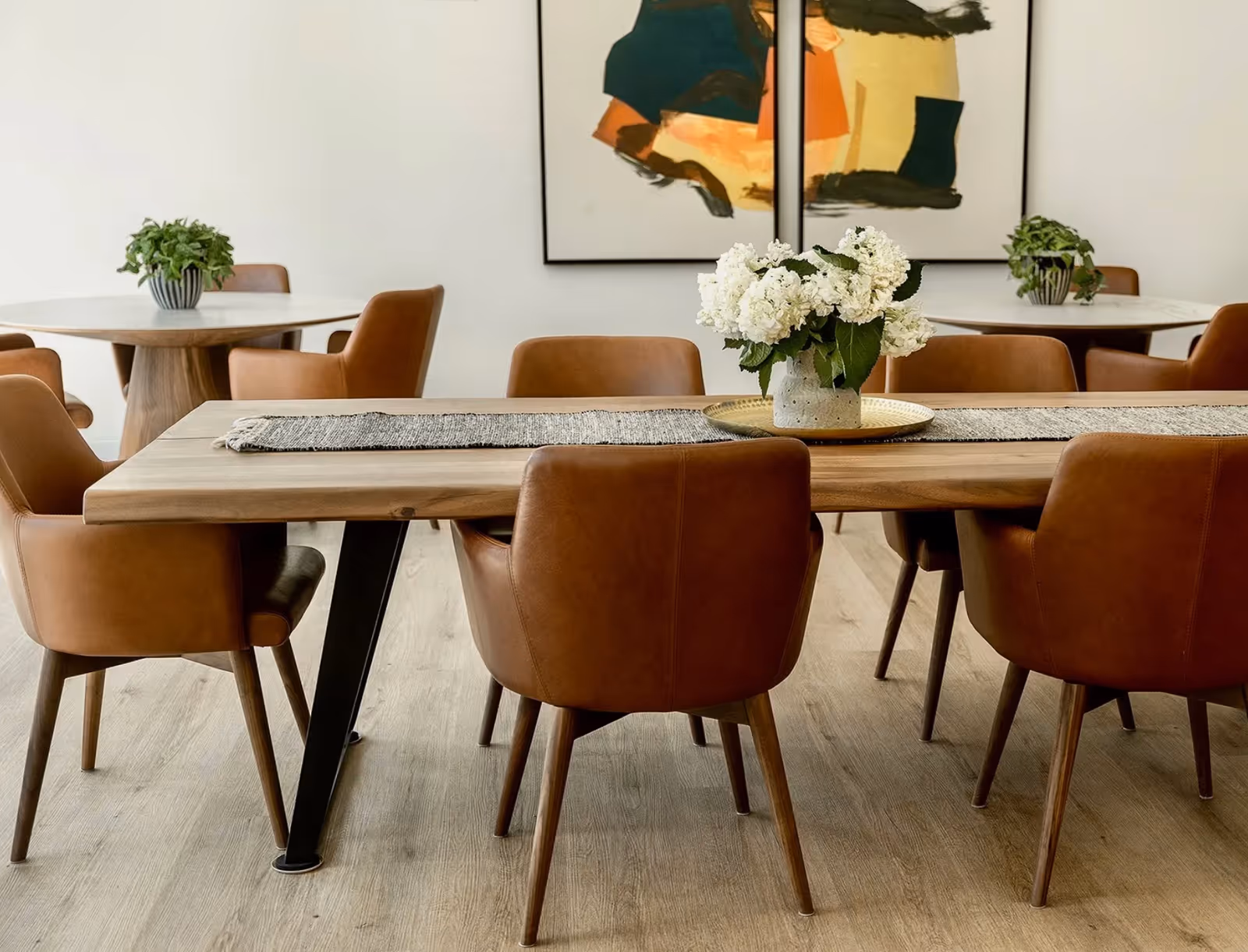 A modern dining room with a wooden table surrounded by brown leather chairs. The table has a gray runner and a vase with white flowers on a golden tray. In the background, there are two round tables with similar brown chairs and green potted plants. Abstract artwork is displayed on the white wall behind the tables.