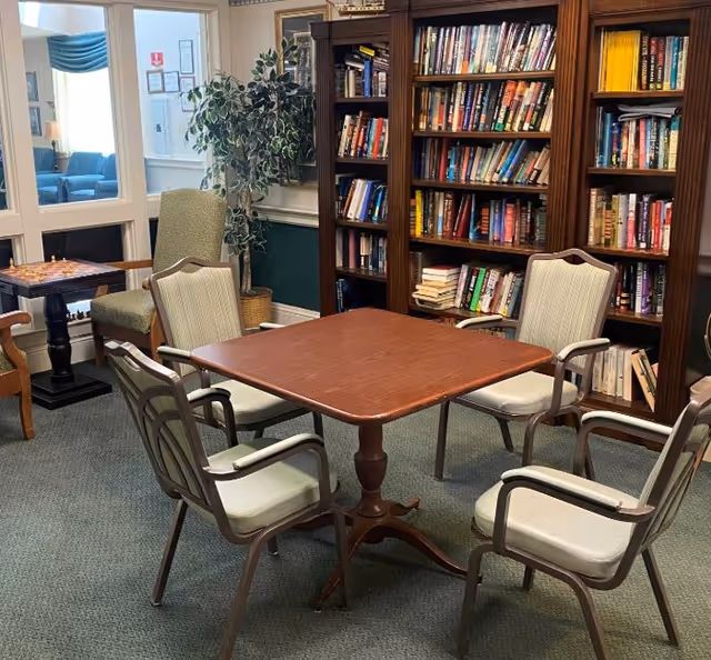 A cozy reading and game area in a senior living facility featuring a square wooden table surrounded by four cushioned chairs. Behind the table is a large wooden bookshelf filled with books. To the left, there is a small table with a chessboard and two armchairs. A potted plant and a window with a view of another seating area are also visible.