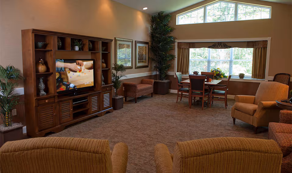 A cozy senior living common room with upholstered chairs, a wooden entertainment cabinet with TV, a small dining table by a large window, and potted plants.