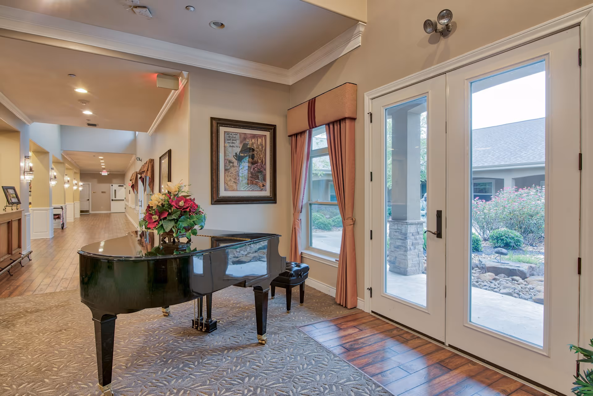Interior view of a senior living facility hallway with a black grand piano adorned with a floral arrangement. The hallway has wooden flooring and beige walls with framed artwork and windows with red curtains. Double glass doors lead to an outdoor garden area with shrubs and a stone pathway.