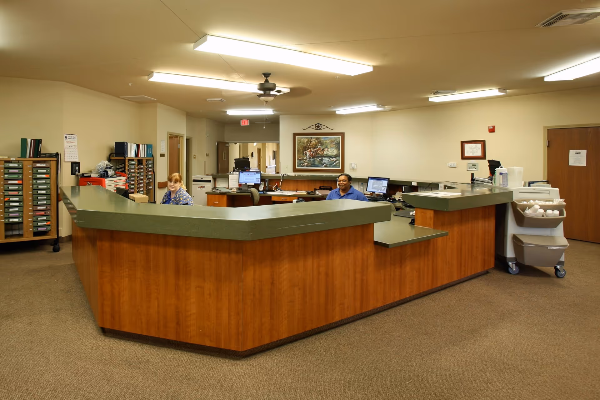 Reception area inside a senior care facility with a large wooden desk and green countertop. Two staff members are seated behind the desk with computer monitors. The room has beige walls, carpeted floor, fluorescent ceiling lights, and a ceiling fan. There are filing cabinets and office supplies visible in the background.