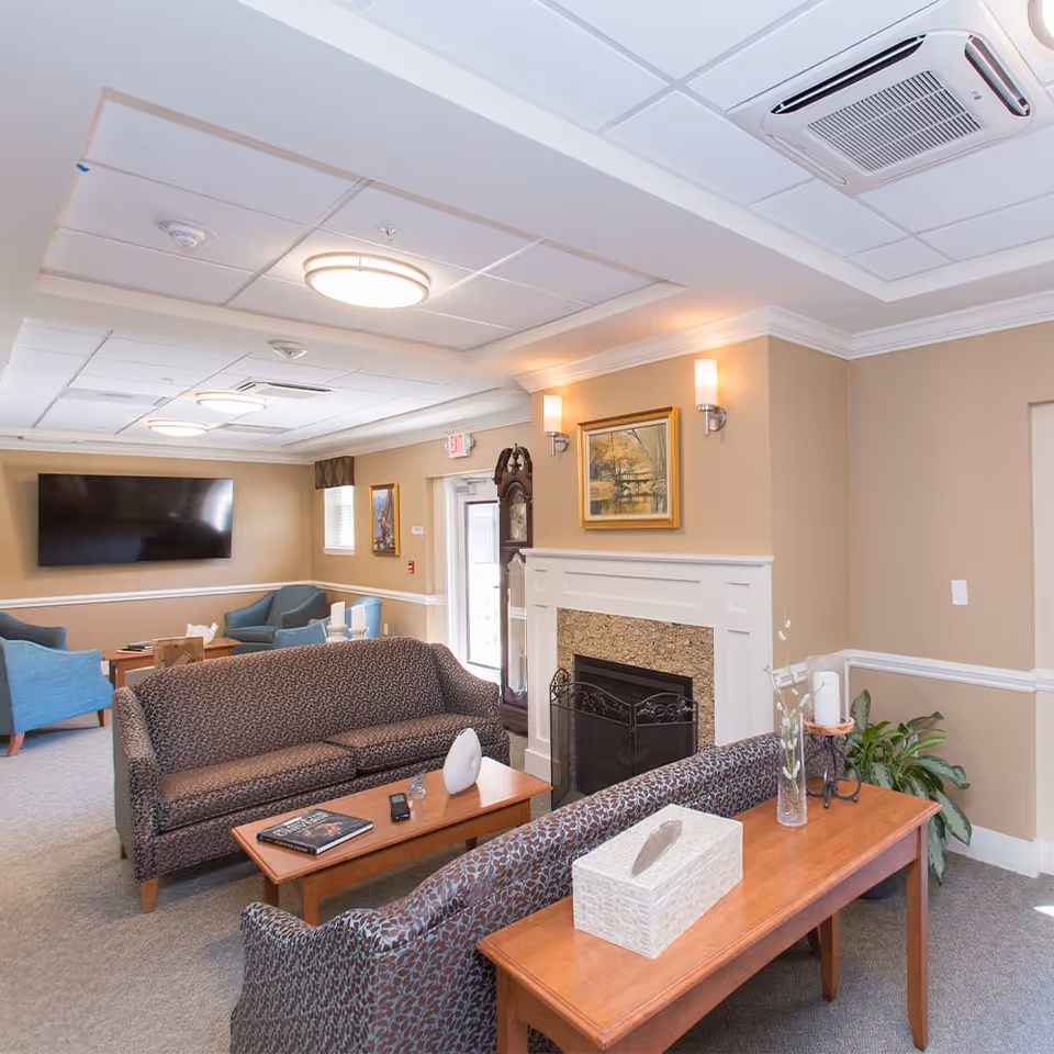 A cozy senior living facility common area with two patterned sofas facing each other, a wooden coffee table with a book and decorative items, a fireplace with a painting above it, a grandfather clock, and a wall-mounted flat screen TV. The room has beige walls, carpeted floor, ceiling lights, and a few blue armchairs near the TV.
