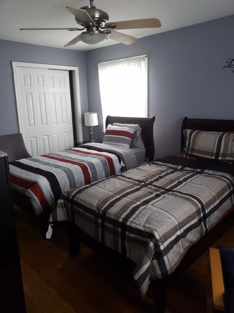 A bedroom with two twin beds, each with different striped bedding. There is a window with sheer curtains between the beds, a ceiling fan above, a lamp on a nightstand, a closet with white sliding doors, and a chair partially visible in the corner.