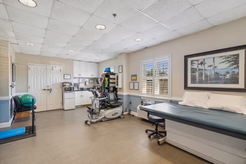 Physical therapy room with a recumbent exercise machine, treatment table, cabinets, and fitness equipment.