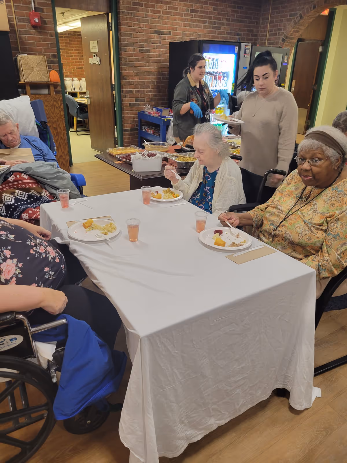 Several elderly individuals seated around a table with a white tablecloth, eating a meal. Two women stand behind the table near a vending machine and a buffet table with food trays. The setting appears to be a communal dining area with brick walls and wooden flooring.