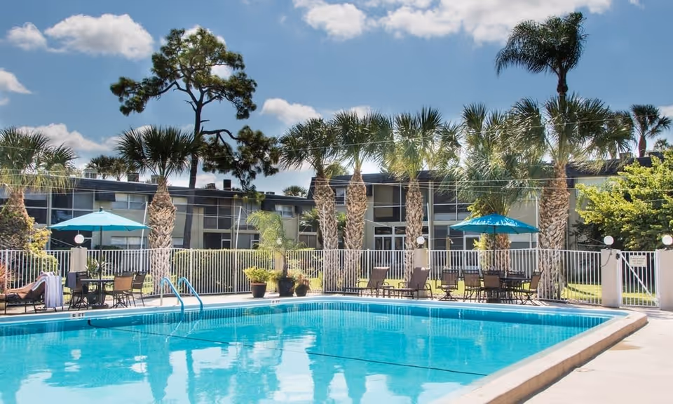 Outdoor swimming pool area at a senior living facility with clear blue water, surrounded by a white fence, palm trees, patio tables with umbrellas, and lounge chairs under a partly cloudy sky.