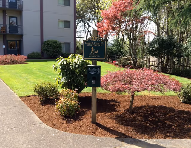Outdoor landscaped area with a small tree, bushes, and a signpost that reads 'Leash & Clean Up after your pet. It's the Law $15 to $200 Fine' with a pet waste bag dispenser attached. A building with balconies and windows is visible in the background.