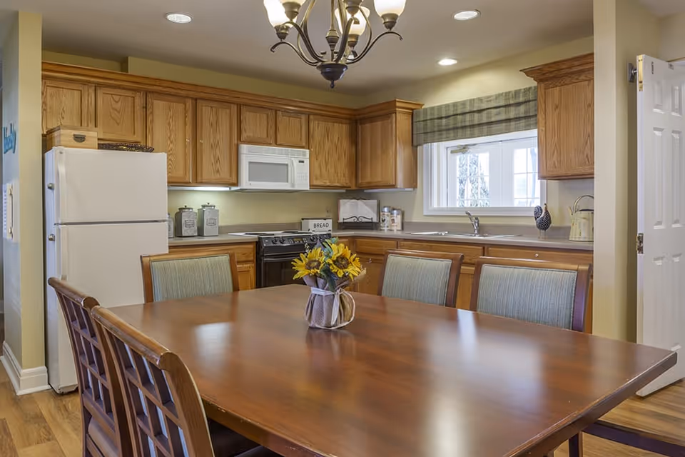 A kitchen and dining area with wooden cabinets, a white refrigerator, a microwave above a stove, and a wooden dining table with six chairs. A vase with sunflowers is placed on the table. There is a window with a plaid valance above the sink, and a chandelier light fixture hangs from the ceiling.