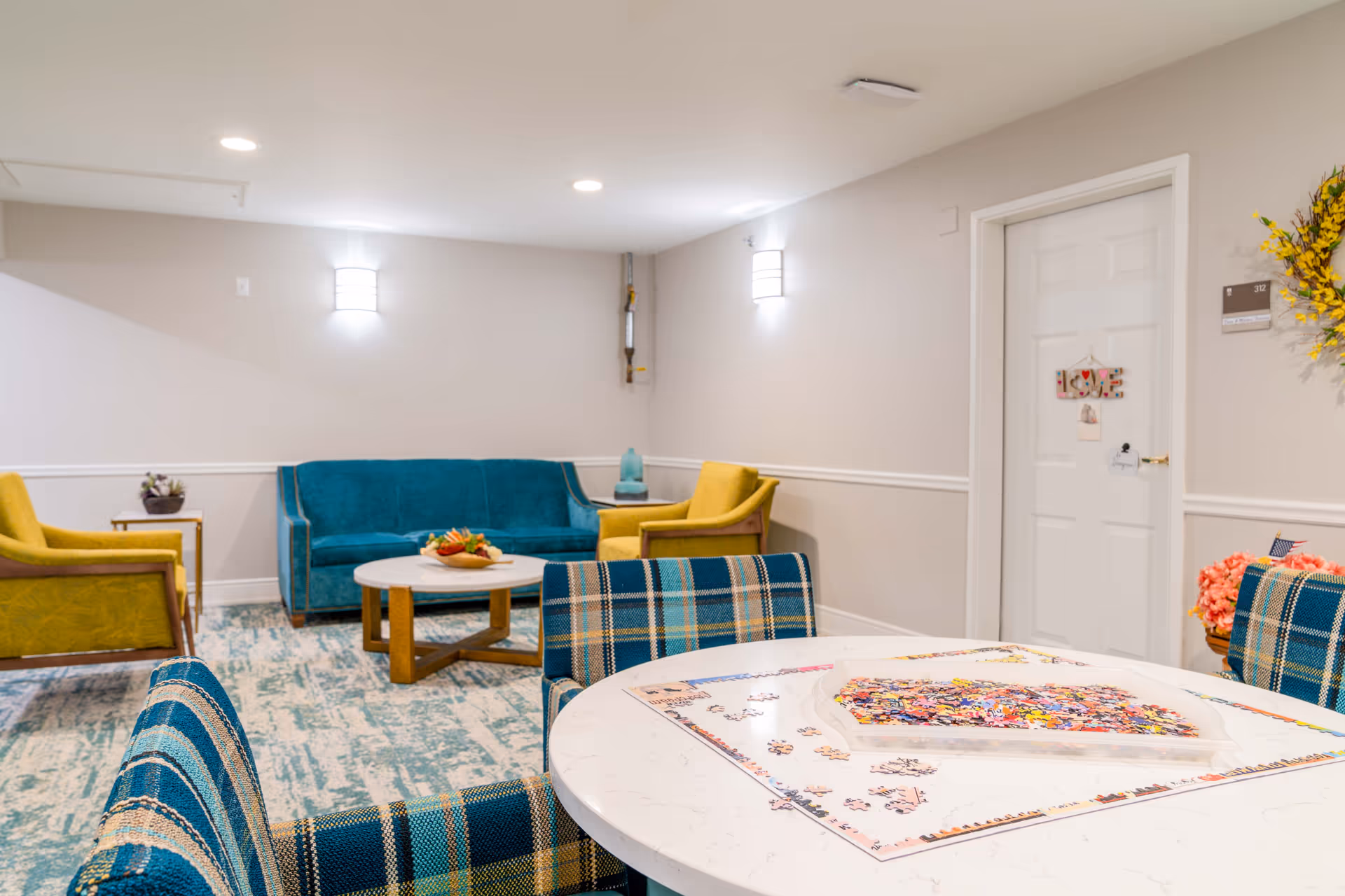 A bright communal living area with a teal sofa, yellow armchairs, plaid dining chairs around a round table with a jigsaw puzzle, and a coffee table.