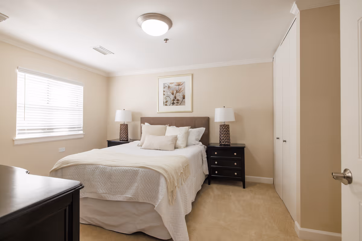 Neutral, well-lit bedroom with a made bed, two bedside tables and lamps, a framed picture above the headboard, and a window with blinds.