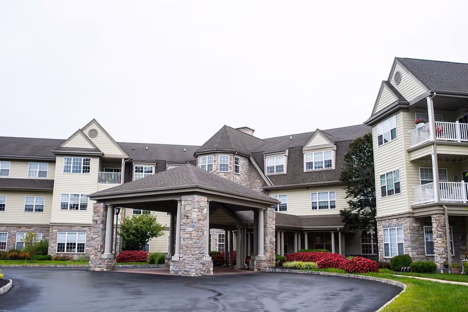 Exterior view of a multi-story senior living facility with beige siding and stone accents. The building features multiple windows, balconies with white railings, and a covered entrance supported by stone pillars. There are landscaped bushes and flowers around the entrance and a paved driveway in front.