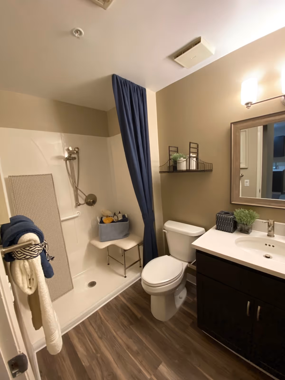 A bathroom with a walk-in shower featuring a handheld showerhead and a white shower bench. There is a blue shower curtain partially drawn. Next to the shower is a toilet, and beside it is a dark wood vanity with a white countertop and an oval sink. Above the vanity is a rectangular mirror with a wooden frame and a wall-mounted light fixture with two bulbs. A small black shelf above the toilet holds decorative items including a small plant. The floor has wood-style vinyl flooring, and a towel rack on the door holds white and blue towels.