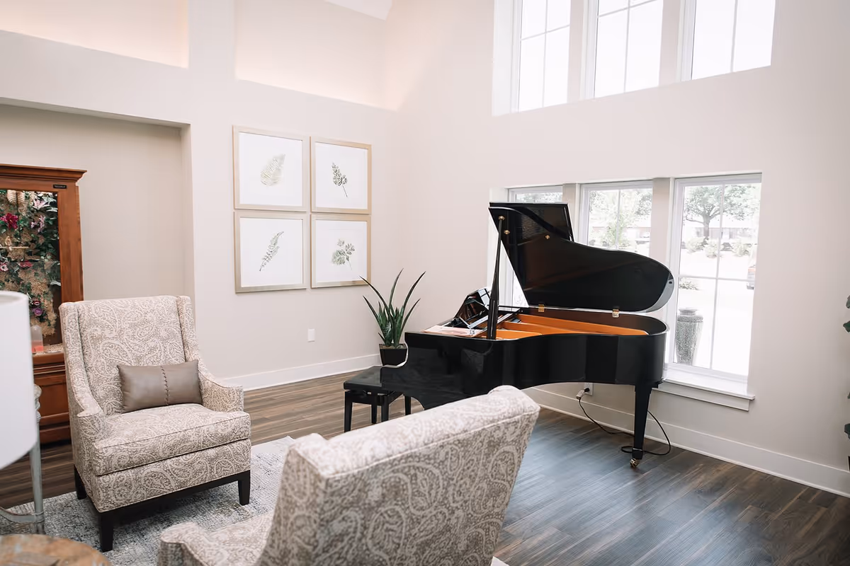 Bright sitting room with a black grand piano by tall windows, patterned armchairs, framed botanical prints, and a potted plant.