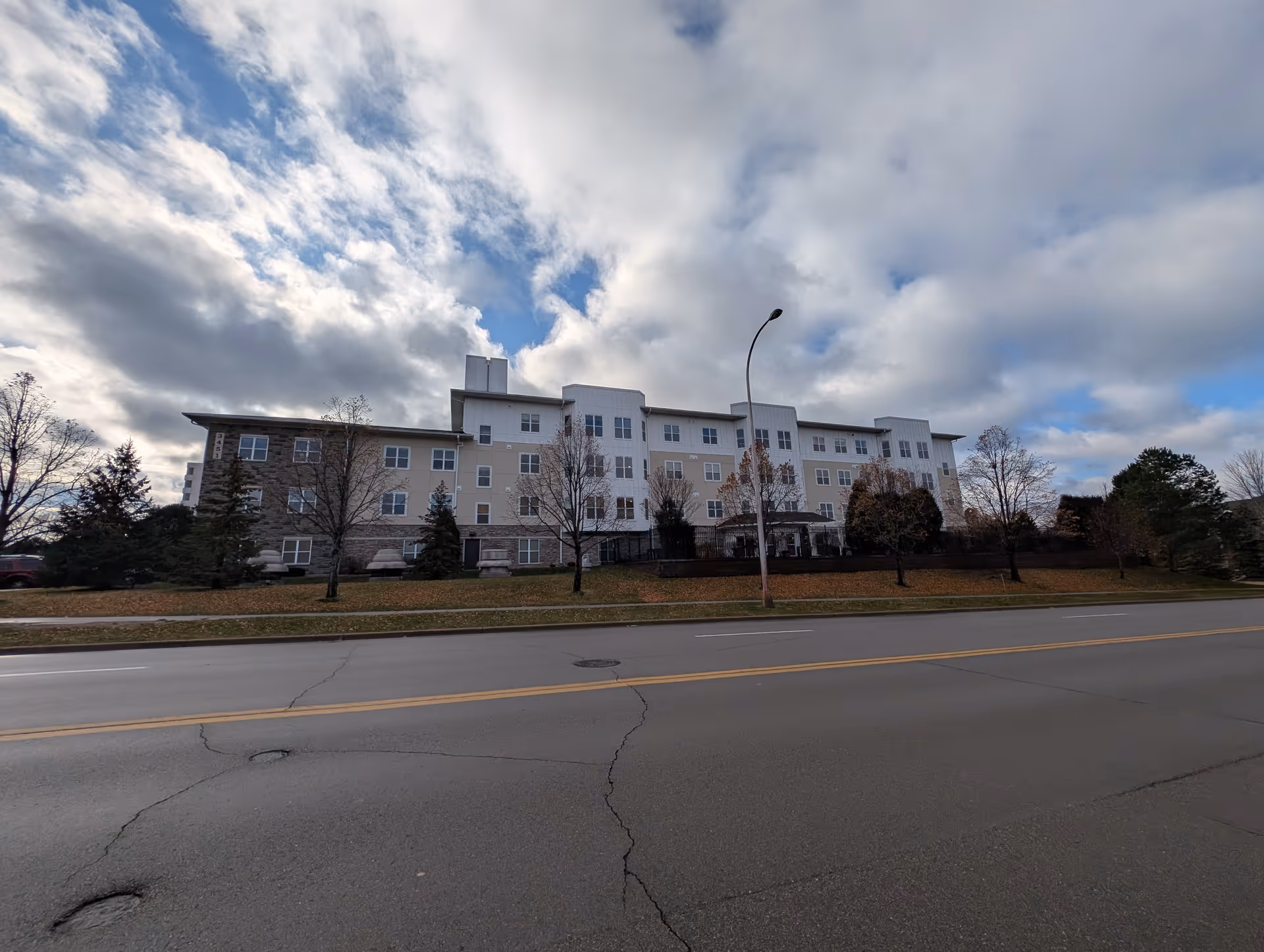 Four-story senior living building with white and beige facade along a street under a cloudy sky.