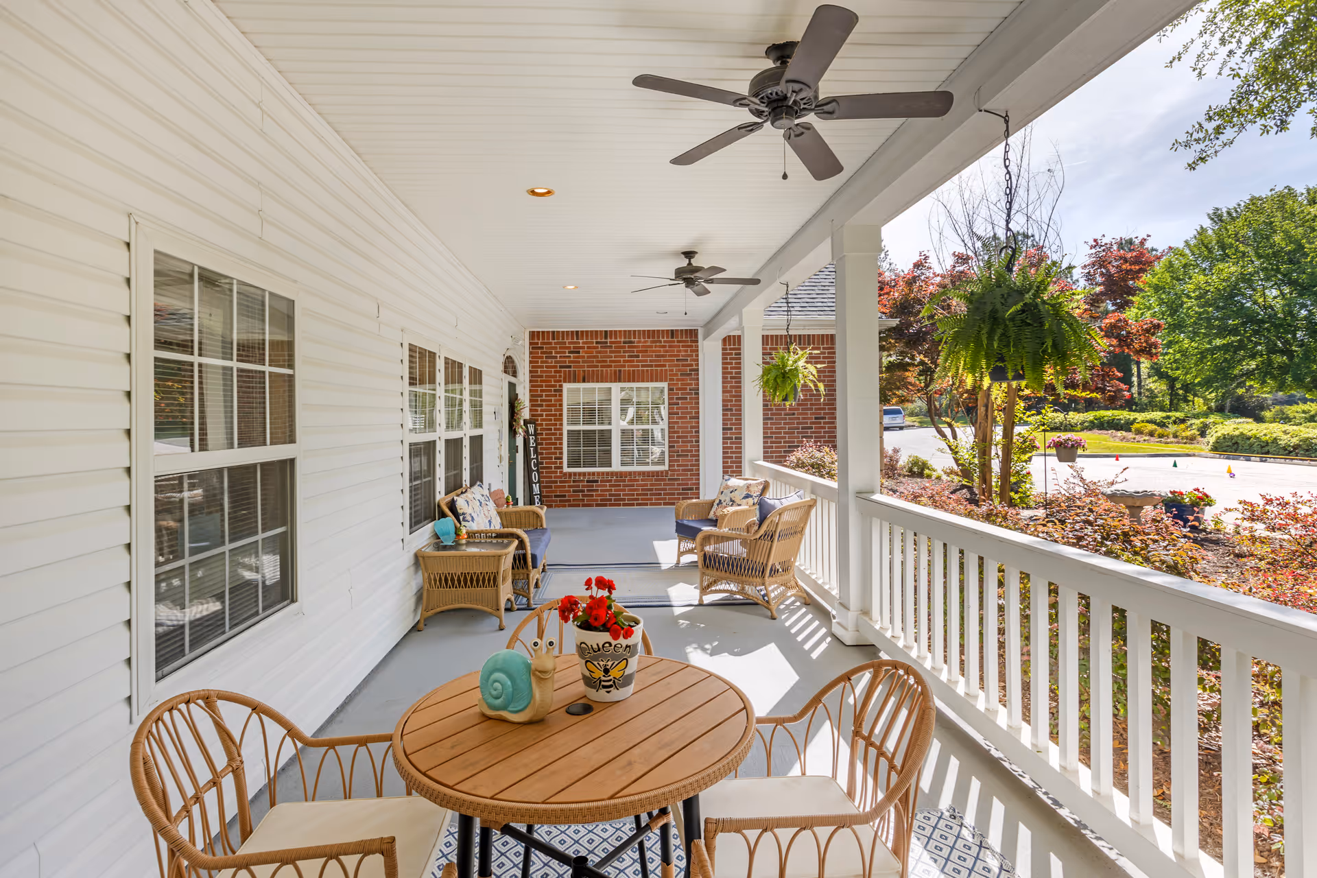 Covered front porch with wicker chairs, a round table, ceiling fans and hanging ferns overlooking landscaped grounds.