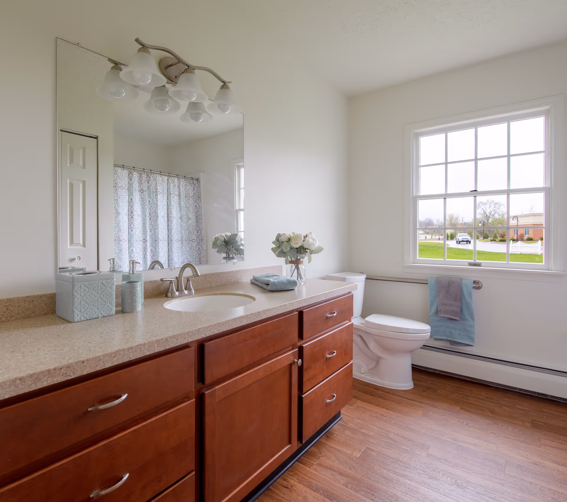Bright bathroom with a large mirror above a beige countertop with wooden cabinets. The bathroom features a sink, a toilet, a window with a view outside, a towel rack with a blue and gray towel, and a shower with a patterned curtain.