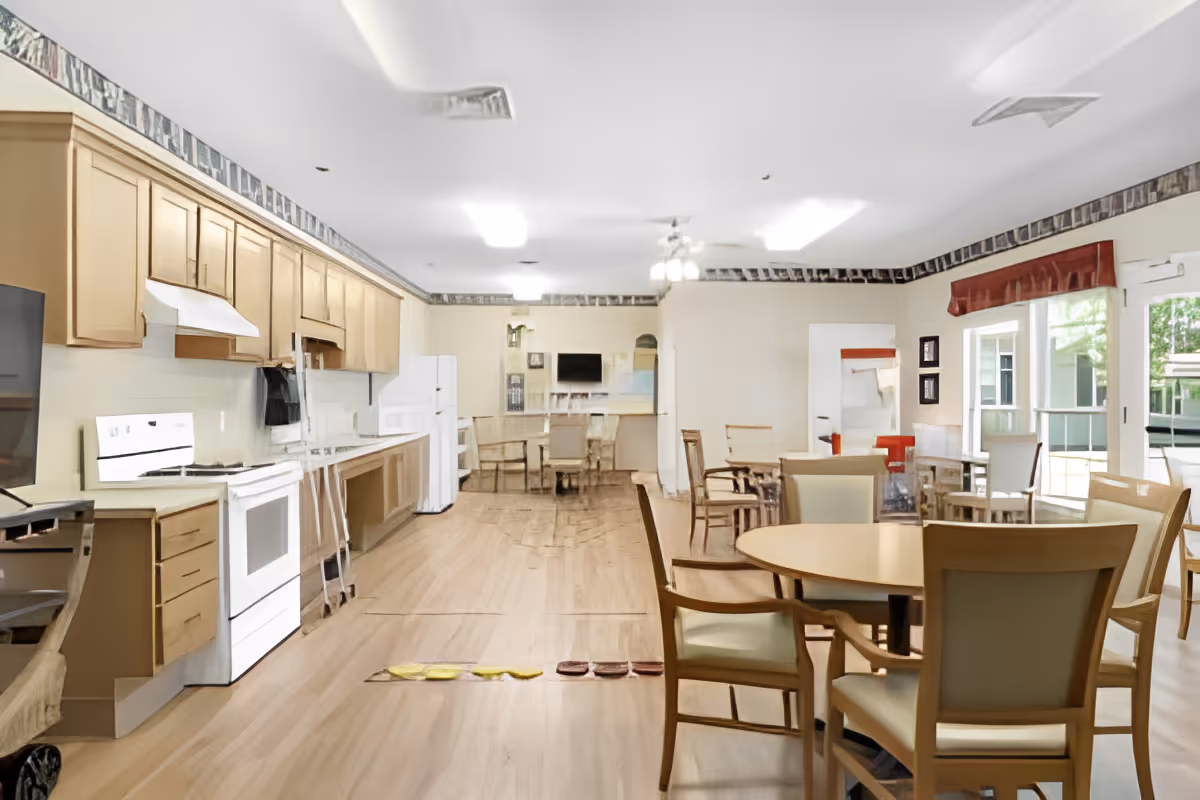 Open communal dining area with a kitchenette along one wall, round tables and chairs, and large windows.