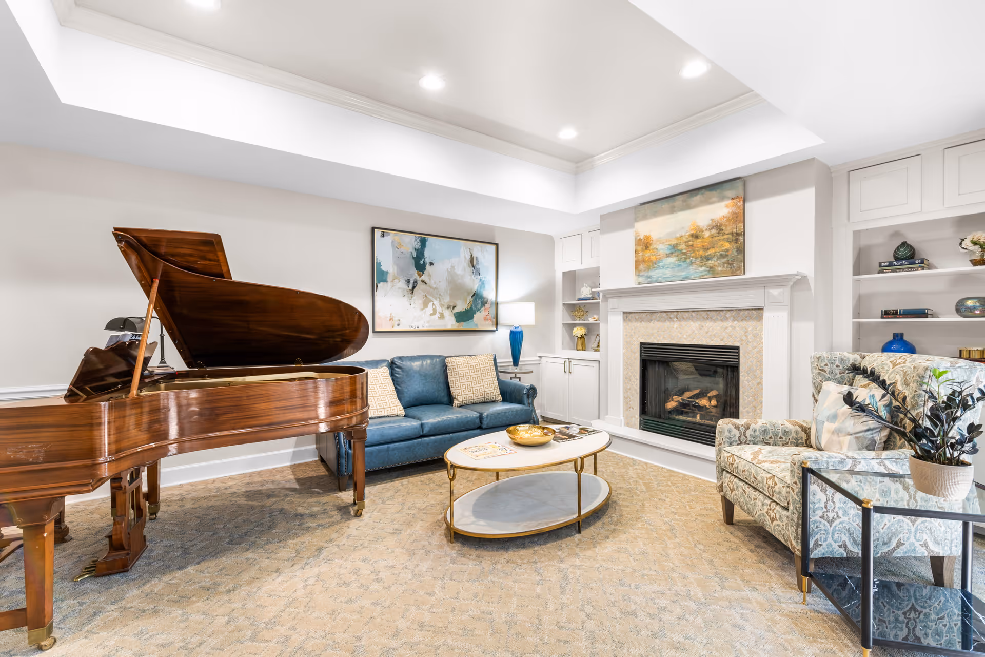 A bright and elegant living room featuring a grand piano on the left, a blue sofa with two patterned cushions, a round coffee table with a gold bowl and magazines, a fireplace with a landscape painting above it, and a patterned armchair with a side table holding a potted plant. Built-in shelves with decorative items flank the fireplace.