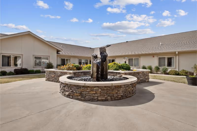 Outdoor courtyard area of a senior living facility with a circular stone water fountain in the center, surrounded by a concrete walkway and landscaped with bushes and flowers near the building. The building has beige walls and multiple windows under a sloped roof, with a partly cloudy blue sky above.