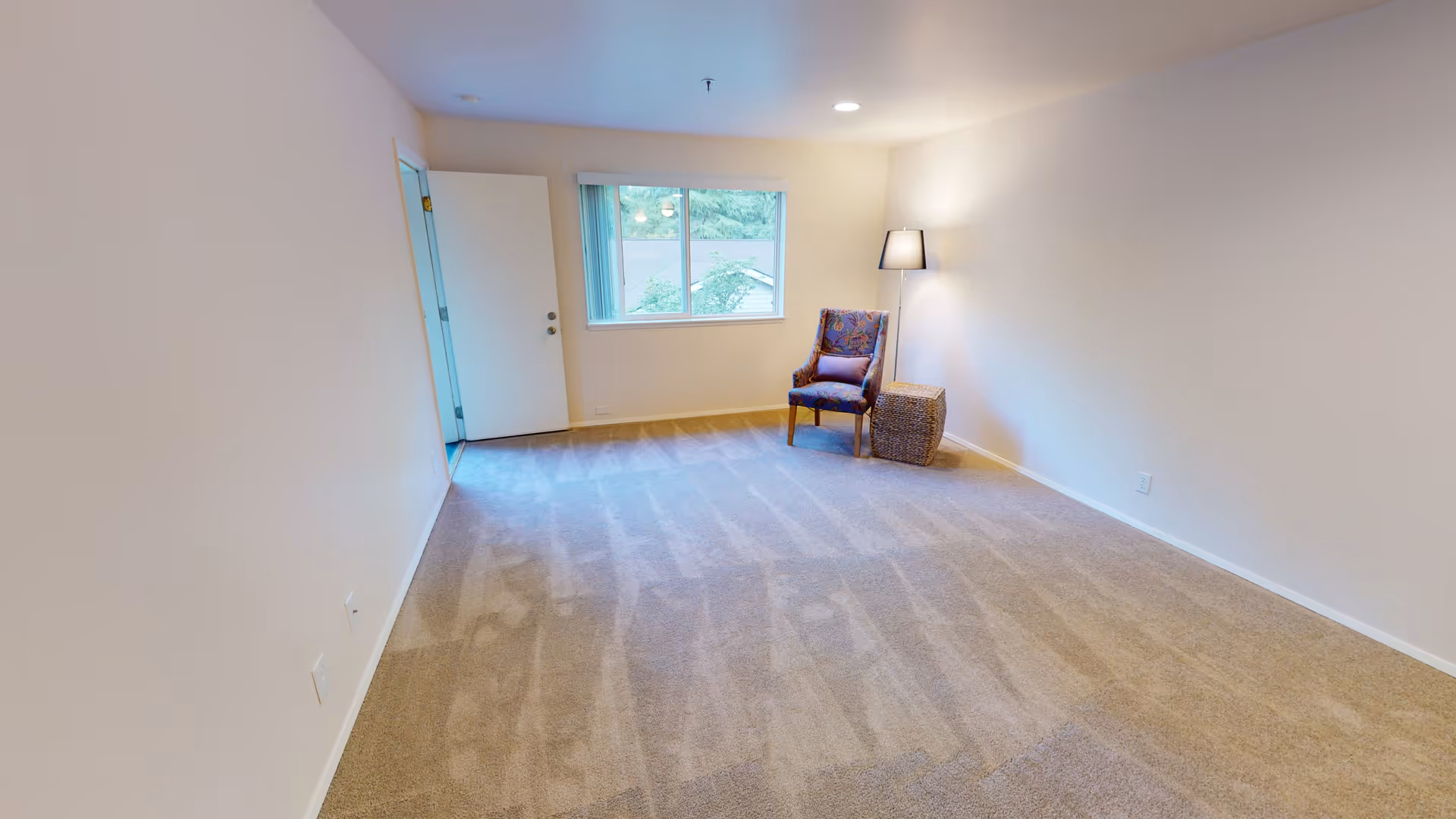 A spacious, empty room with beige carpet and white walls, featuring a single upholstered chair with a cushion and a floor lamp next to a woven basket near a window with a view of greenery outside. There is an open door leading to another room.