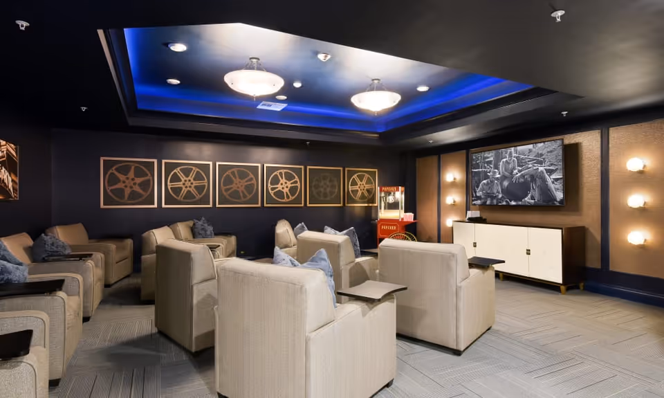 A cozy movie theater room with beige armchairs arranged in rows facing a large flat-screen TV mounted on the wall. The walls are decorated with framed images of film reels and the ceiling has blue accent lighting with two ceiling lights. A popcorn machine is visible in the corner of the room.