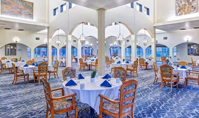A spacious and elegant dining room with round tables covered in white tablecloths, each set with blue napkins, white mugs, and small potted plants. The room features high ceilings with large arched openings and modern chandeliers. The carpet has a blue and white pattern, and the walls are decorated with large paintings.