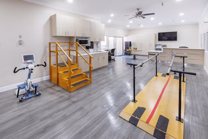 A spacious rehabilitation room with wooden parallel bars on a yellow platform for walking exercises, a small set of wooden stairs with handrails, an exercise bike, and a kitchenette area with cabinets, microwave, and sink. The room has gray flooring, white walls, a ceiling fan, and a wall-mounted TV.