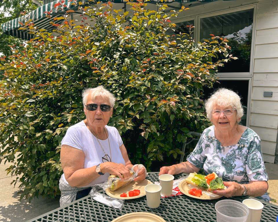 Two elderly women sitting at a green metal outdoor table enjoying a meal with sandwiches and watermelon slices. They are seated in front of a large leafy bush with a building wall and window behind them. One woman is wearing sunglasses and a white shirt, while the other is wearing glasses and a white shirt with a green floral pattern.