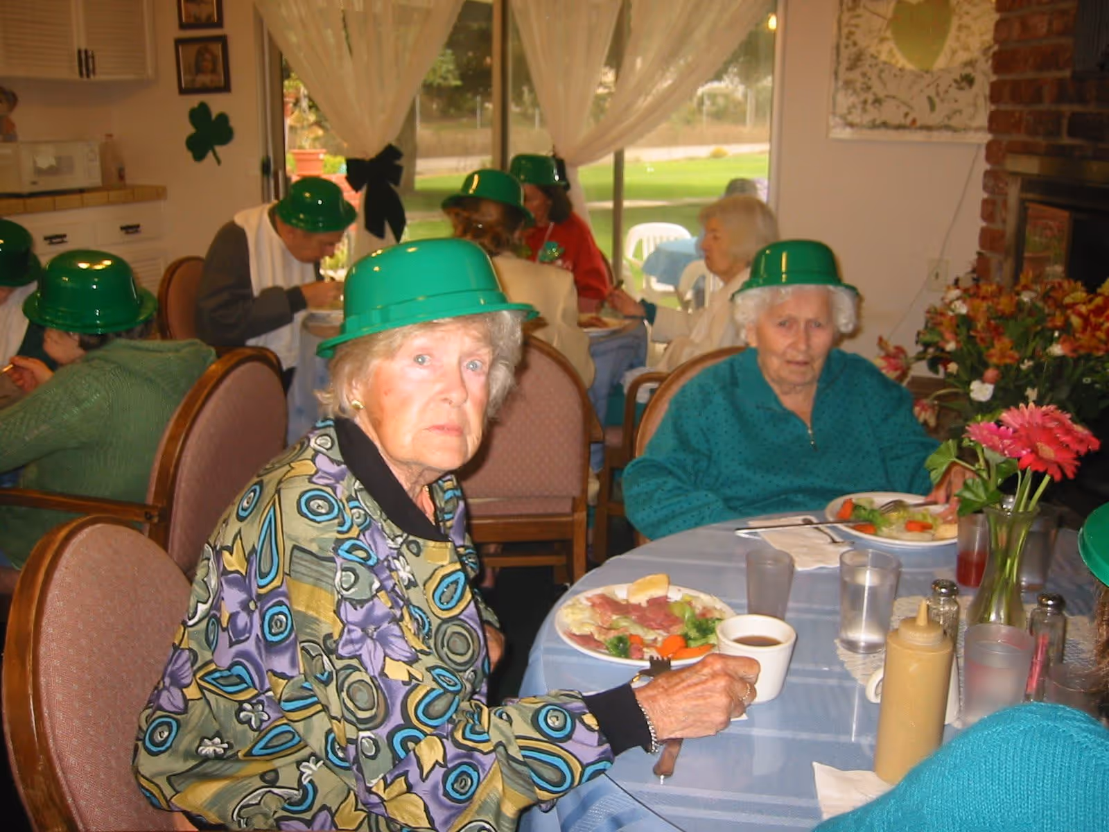 A group of elderly people wearing green hats are seated around tables in a dining area, eating a meal. The room has large windows with sheer curtains, and there are flowers on the table. The atmosphere suggests a social gathering or celebration.