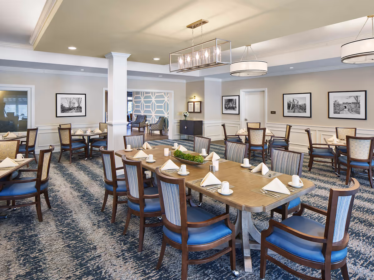 A spacious dining room with multiple wooden tables and chairs arranged neatly. Each table is set with white napkins, cups, and silverware. The room has patterned carpet flooring, light-colored walls adorned with black and white framed photographs, and modern ceiling light fixtures. There is an open doorway leading to another seating area with armchairs and a decorative wall.