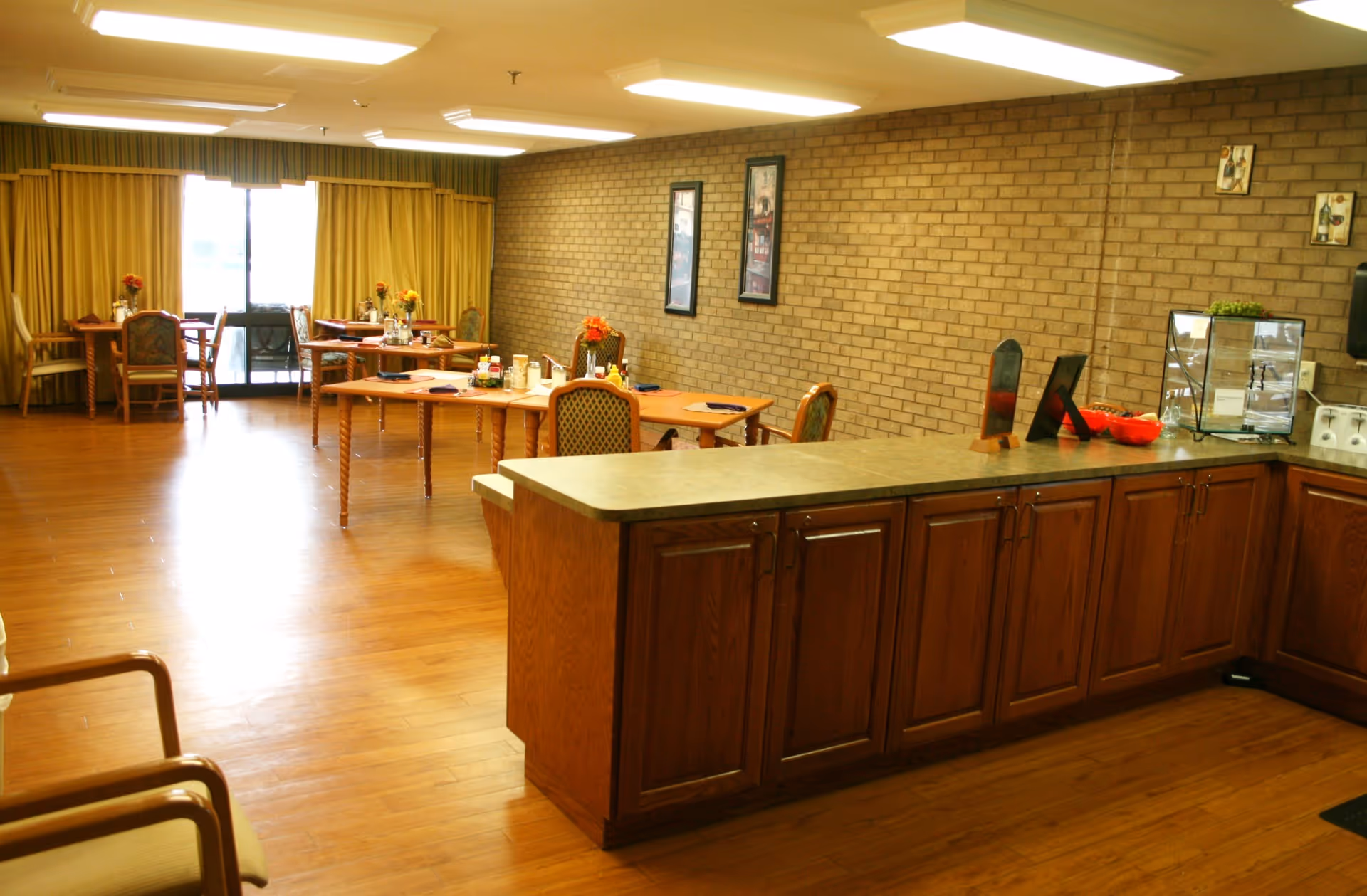 A dining area with wooden floors and a brick wall. Several tables with chairs are set up, each table decorated with a small flower arrangement and place settings. There is a counter with wooden cabinets in the foreground, and large windows with yellow curtains in the background letting in natural light.