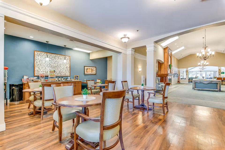 A bright and spacious common area in a senior living facility featuring several round wooden tables with cushioned chairs. The room has wooden flooring and a blue accent wall with framed artwork. Pendant lights hang above a counter area with snacks and a coffee station. In the background, there is a carpeted lounge area with chandeliers and large windows allowing natural light to fill the space.