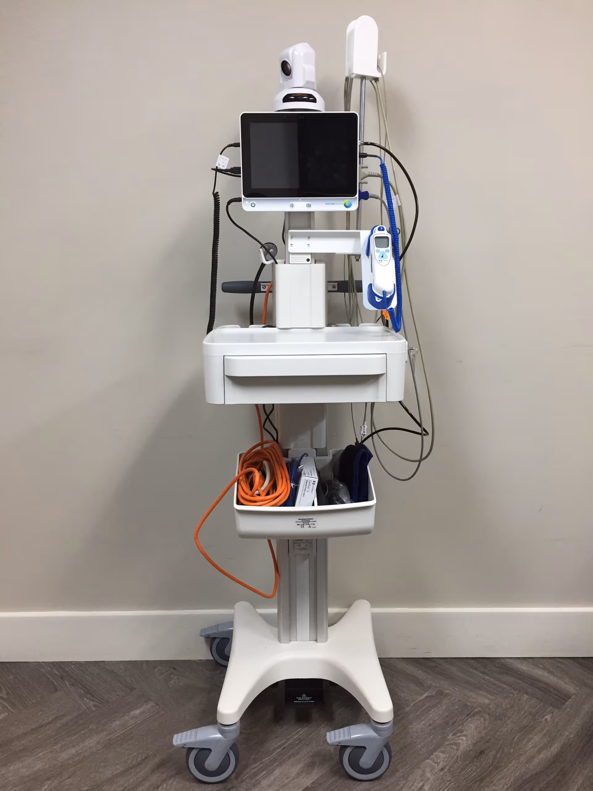 A mobile medical cart with a monitor, various cables, and medical devices attached, positioned against a plain beige wall on a wood-patterned floor.
