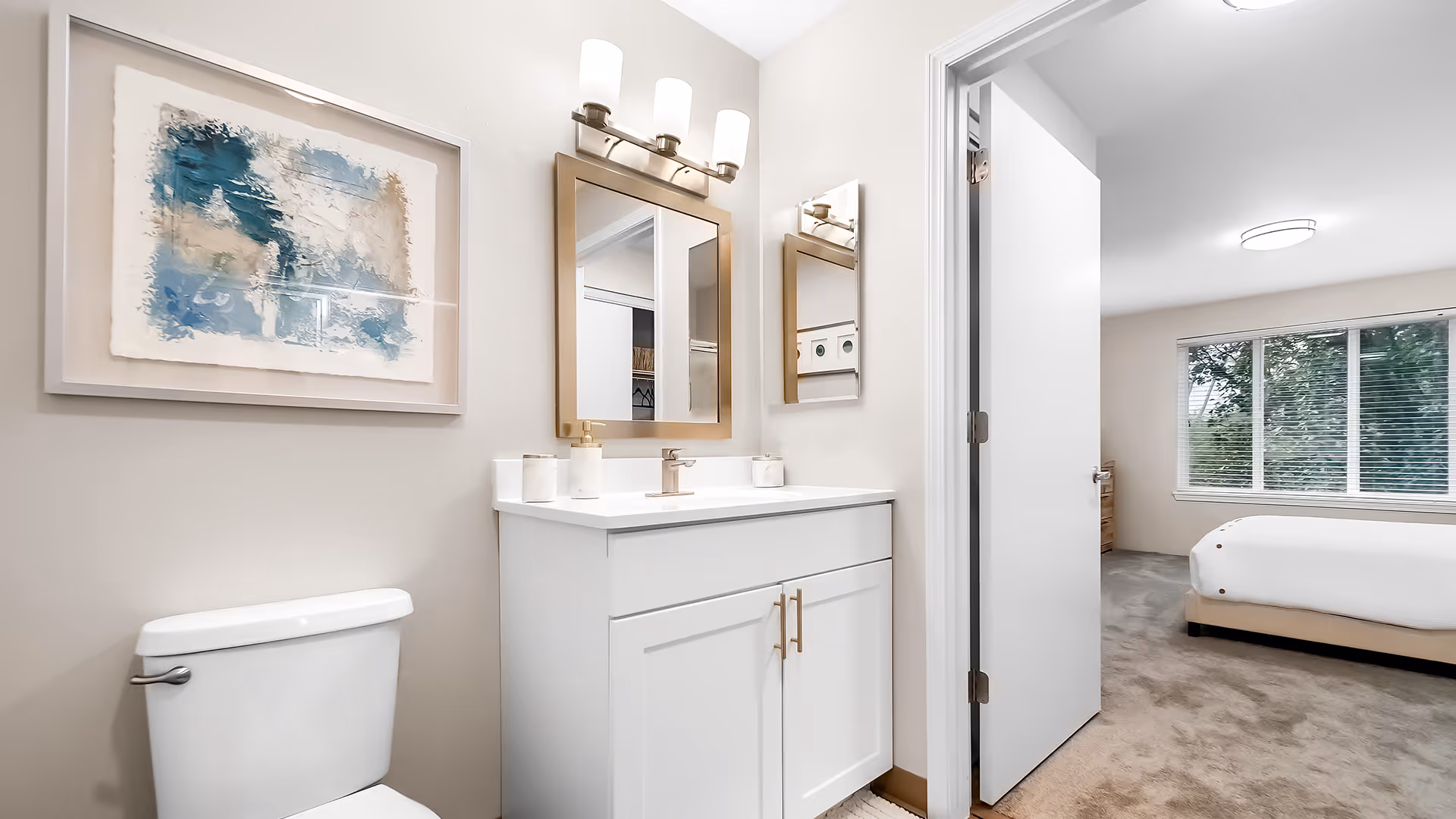 A clean and modern bathroom featuring a white toilet, a white vanity with a sink, a rectangular mirror with a light fixture above it, and abstract artwork on the wall. The bathroom opens into a bedroom with a bed, window with blinds, and carpeted floor.
