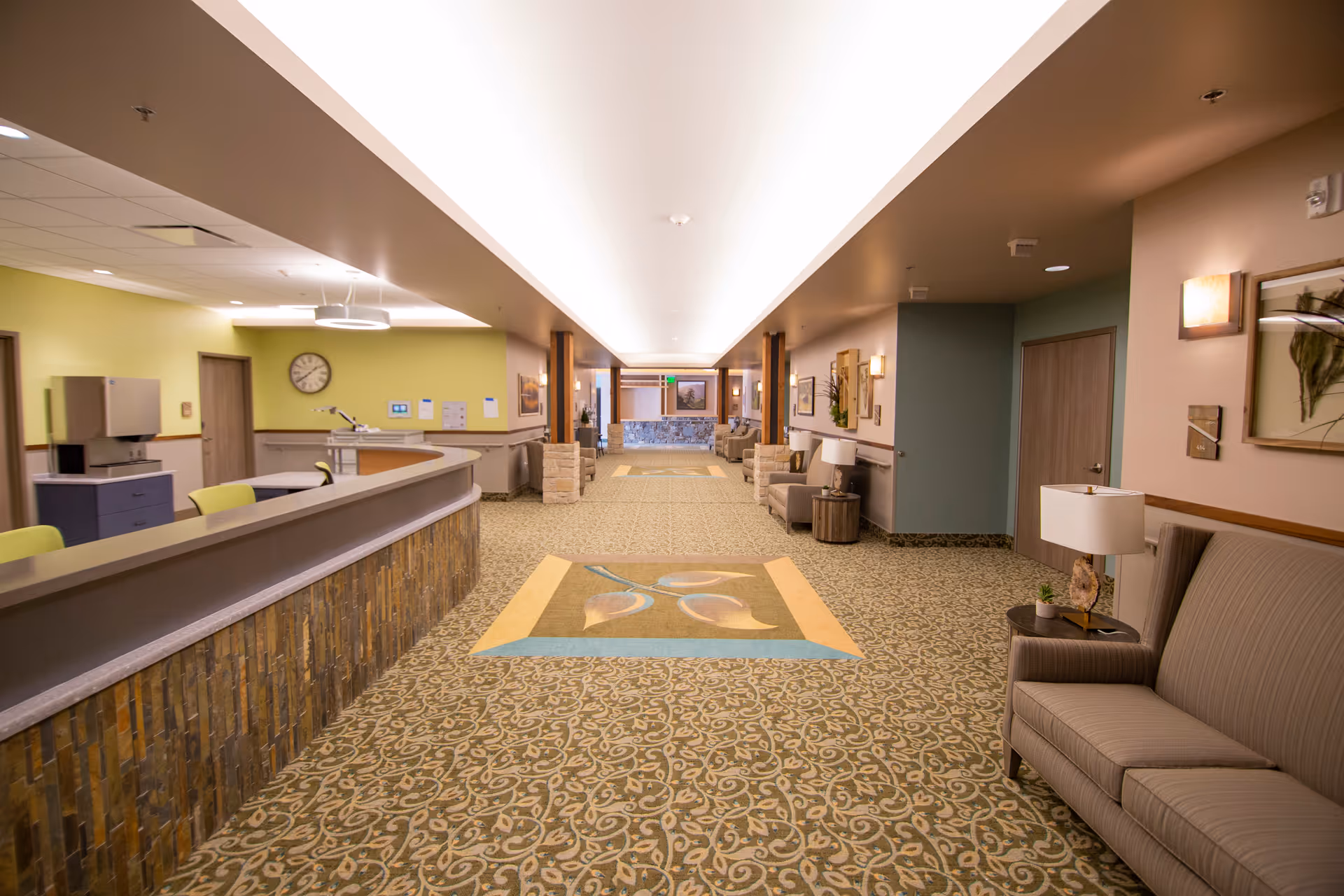 A spacious, well-lit hallway in a senior living facility with a patterned carpet and a decorative rug in the center. On the left side, there is a curved reception desk with chairs behind it and a clock on the wall. On the right side, there are cushioned benches with side tables and lamps, along with framed artwork on the walls. The hallway extends into a seating area with more chairs and tables in the distance.