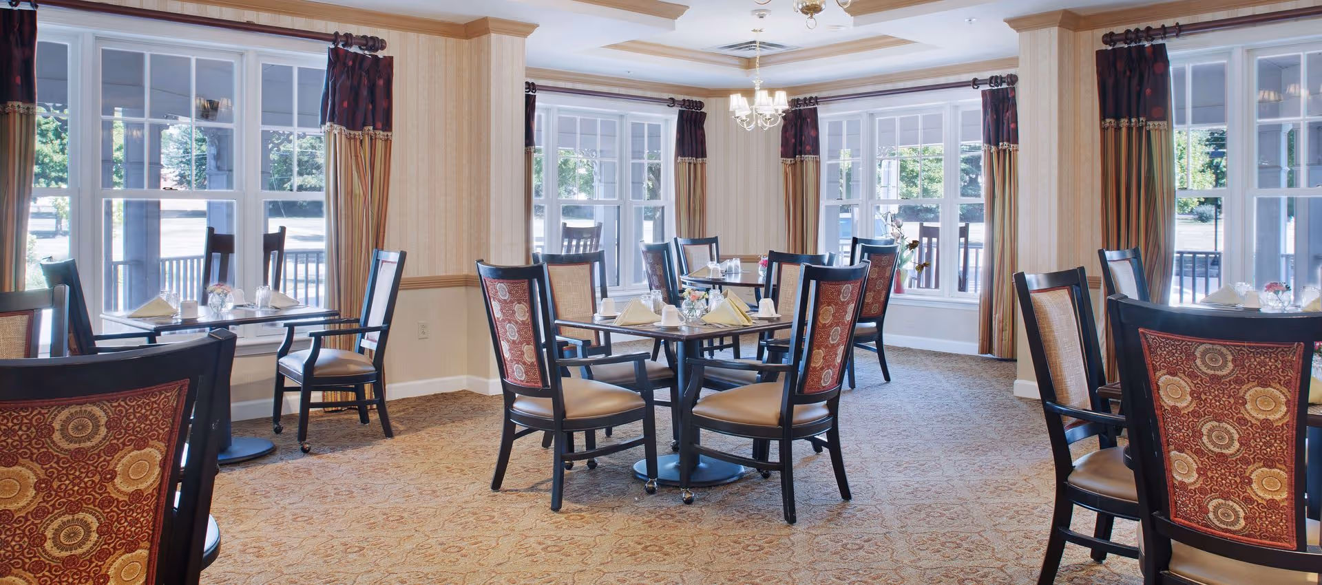 Bright dining room with multiple round tables and patterned chairs set for a meal, large windows and drapery letting in daylight.
