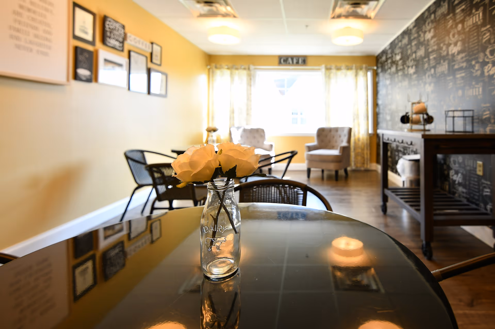 A cozy cafe area with a glass table in the foreground holding a small glass bottle with white flowers. The room has yellow walls with framed pictures and quotes, two cushioned armchairs near a window with yellow curtains, and a dark wooden cart against a wall with black and white cafe-themed wallpaper.
