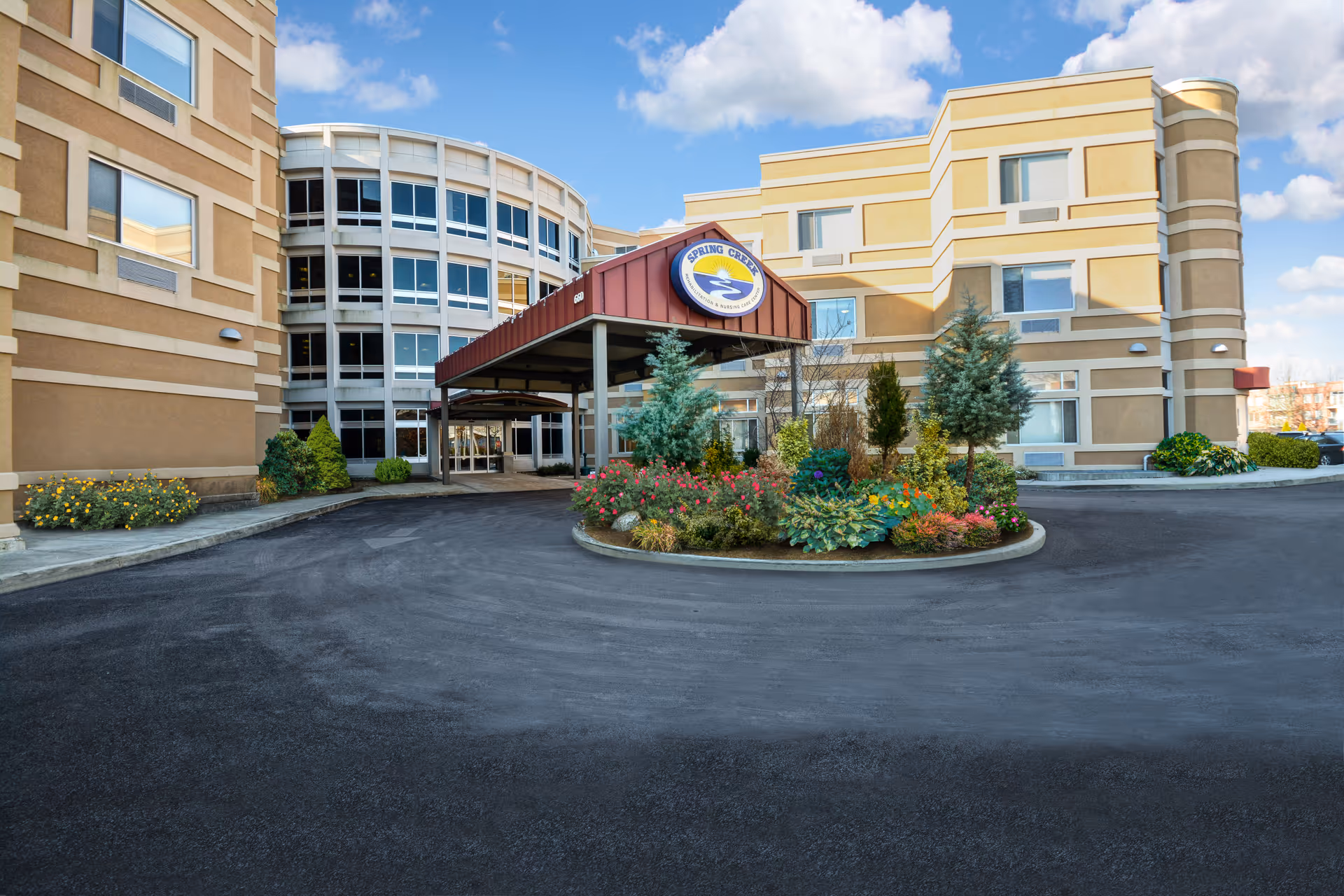 Exterior view of Spring Creek Rehabilitation and Nursing Care Center showing a multi-story building with beige and white walls, a covered entrance with a sign displaying the facility's name and logo, surrounded by landscaped greenery and flowers under a blue sky with some clouds.