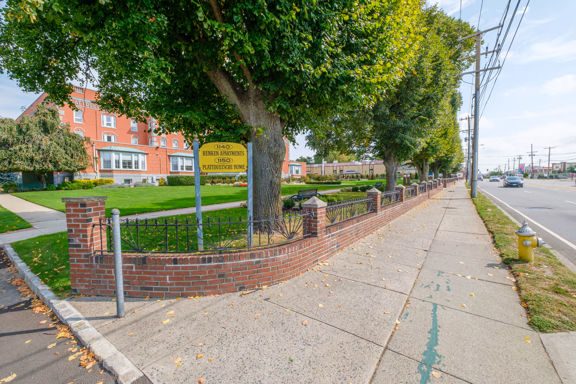 Brick-faced senior housing building with a landscaped front lawn and low brick fence along a sidewalk and street.