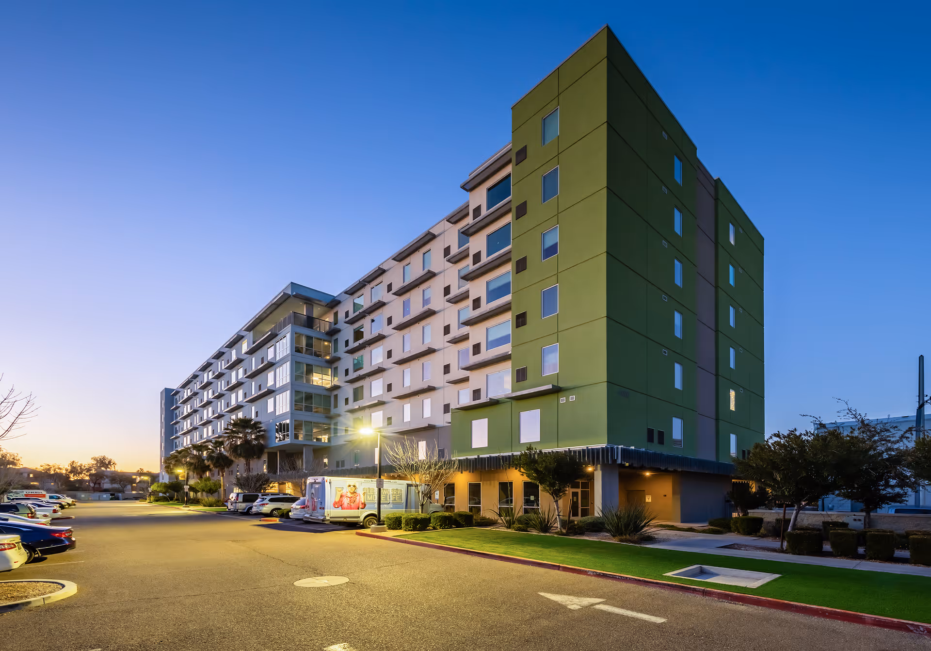 Exterior view of a multi-story senior living facility building at dusk with a parking lot in front. The building has a modern design with green and beige walls and multiple windows. Several cars are parked in the lot, and there are some trees and landscaping around the building.