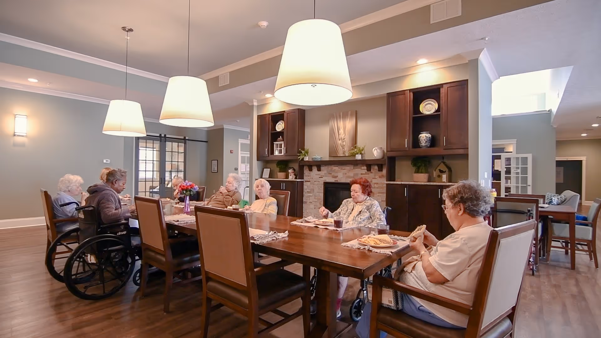 A group of elderly people sitting around a large wooden dining table in a well-lit room with pendant lights hanging from the ceiling. Some individuals are in wheelchairs, and they appear to be eating or socializing. The room has wooden floors, built-in cabinets, and a fireplace with decorative items on the mantel.
