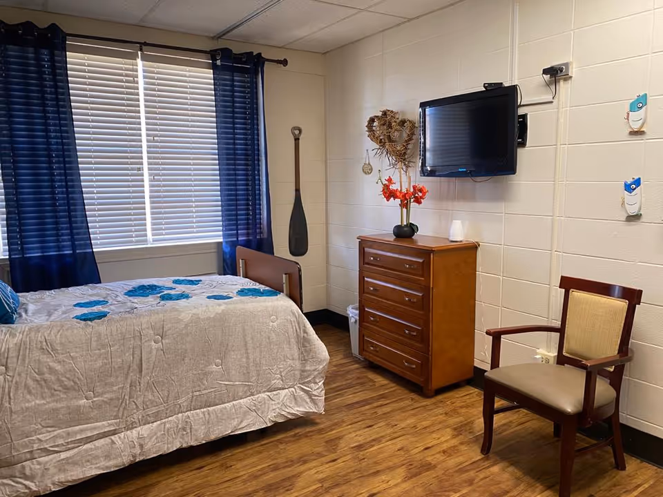 A furnished senior bedroom with a bed, dresser topped with flowers and a wall-mounted TV, a chair, and a window with blue curtains.