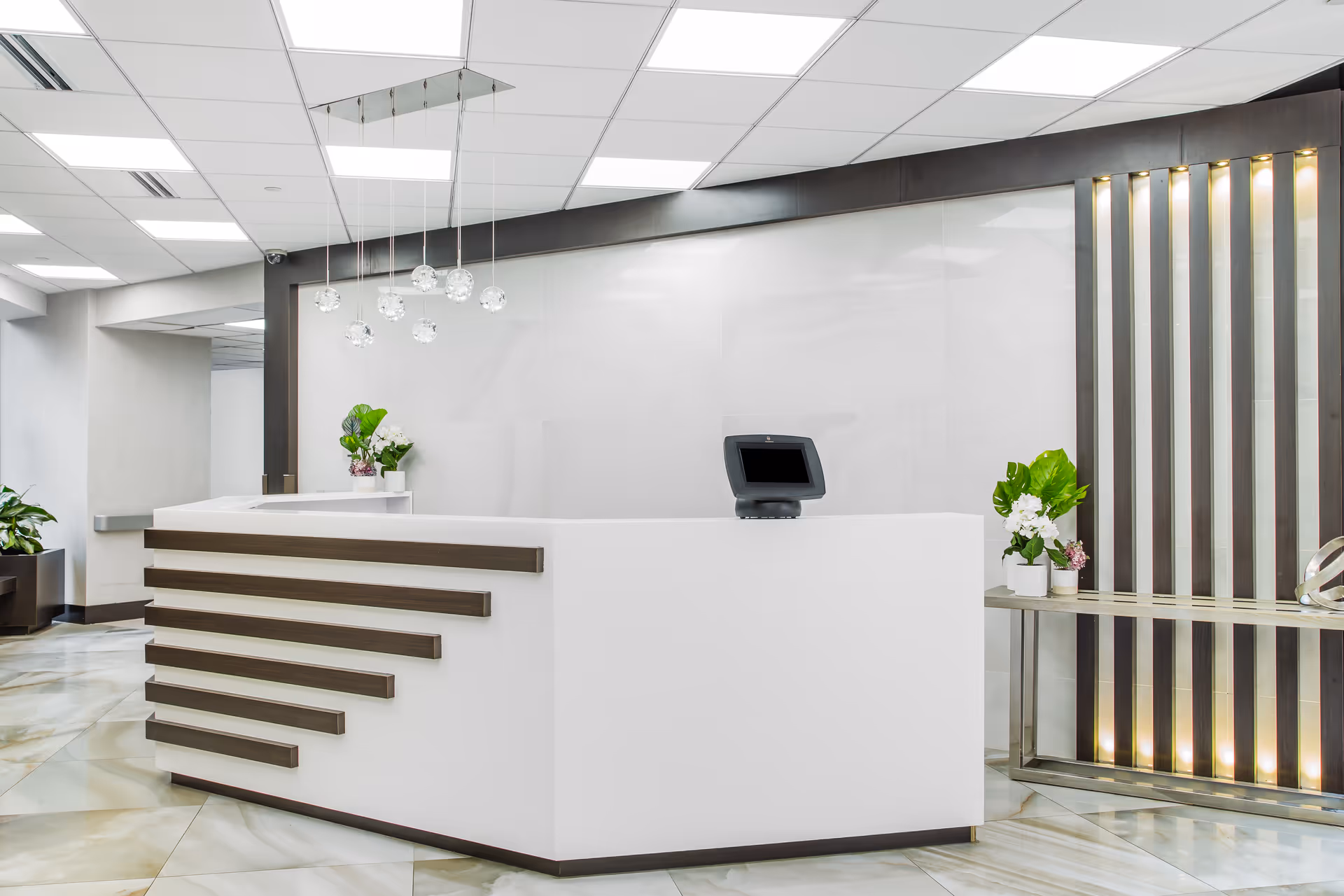 Modern reception desk area with a white counter featuring horizontal dark wood accents, a small monitor on the desk, decorative plants, pendant lights hanging from the ceiling, and a decorative wall with vertical lighted panels behind a side table.