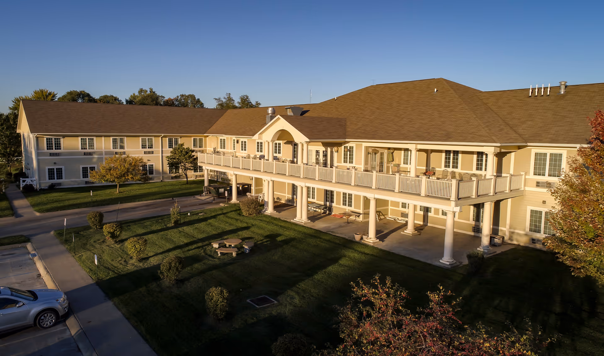 Exterior view of a two-story assisted living facility building with a large balcony supported by columns. The building is surrounded by a well-maintained lawn with small bushes, trees, and a picnic table. A parking area with a silver car is visible on the left side. The sky is clear and blue.