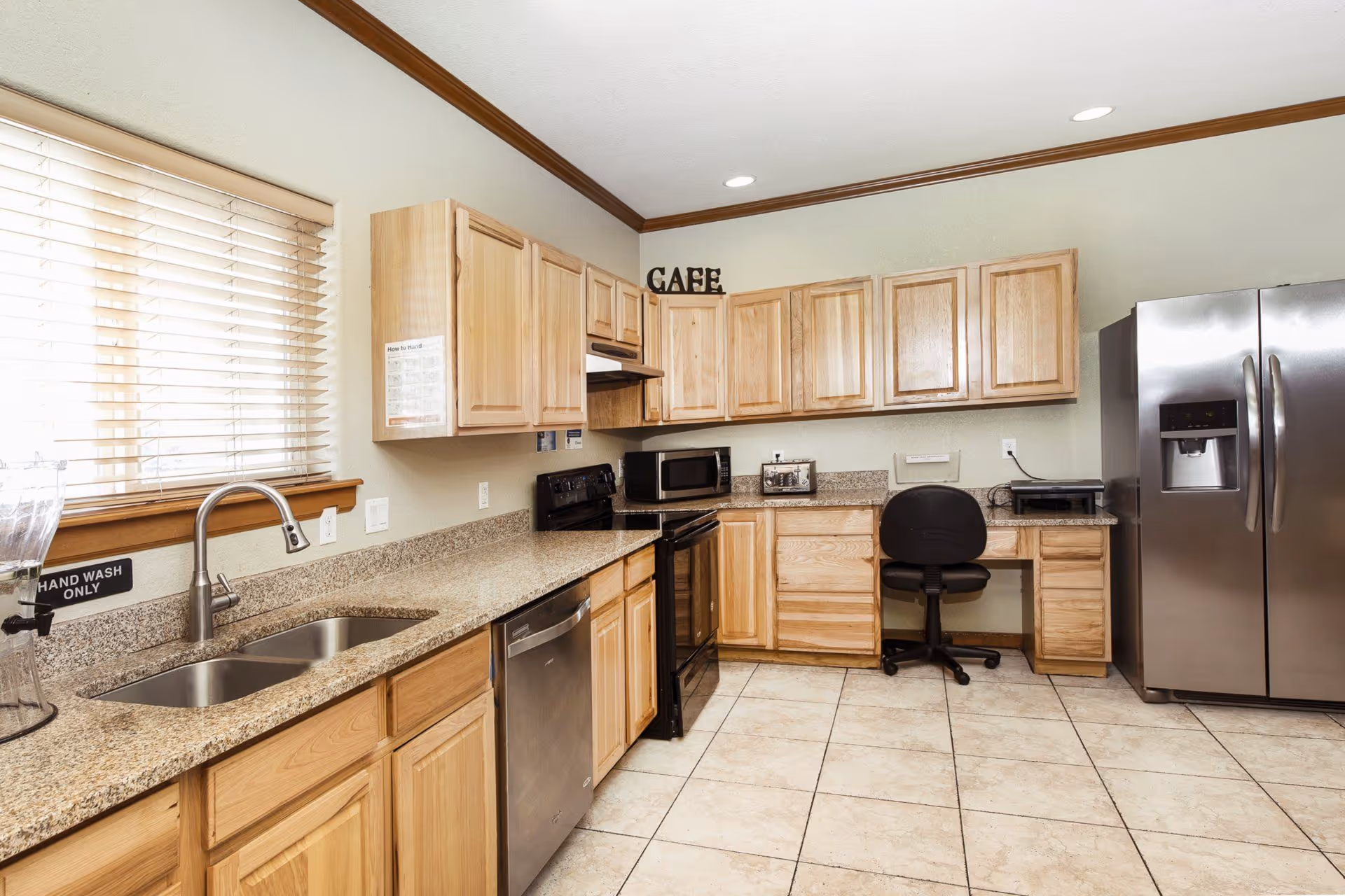 A clean kitchen area with light wood cabinets, granite countertops, and tiled floor. The kitchen features a double sink, dishwasher, black stove, microwave, toaster, and a stainless steel refrigerator. There is a black office chair at a built-in desk area, and a window with blinds letting in natural light. The word 'CAFE' is displayed on top of the cabinets.