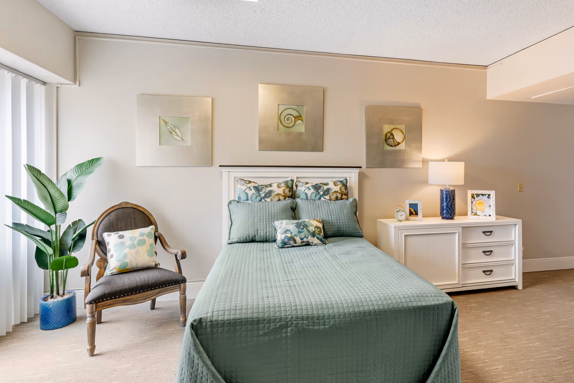 A neatly made bed with a green quilt and floral pillows in a bedroom. To the left of the bed is a wooden chair with a floral cushion and a tall green plant in a blue pot. On the right side of the bed is a white dresser with a blue lamp, two framed pictures, and a small clock. Three square art pieces featuring seashell designs hang on the wall above the bed.