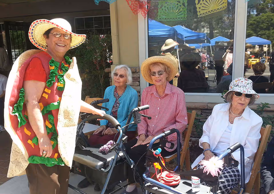 Four elderly women sitting outdoors near a building window, three seated with walkers in front of them and one standing wearing a colorful taco costume and a wide-brimmed hat. The seated women are wearing hats and sunglasses, enjoying a sunny day with blue tents visible in the background.