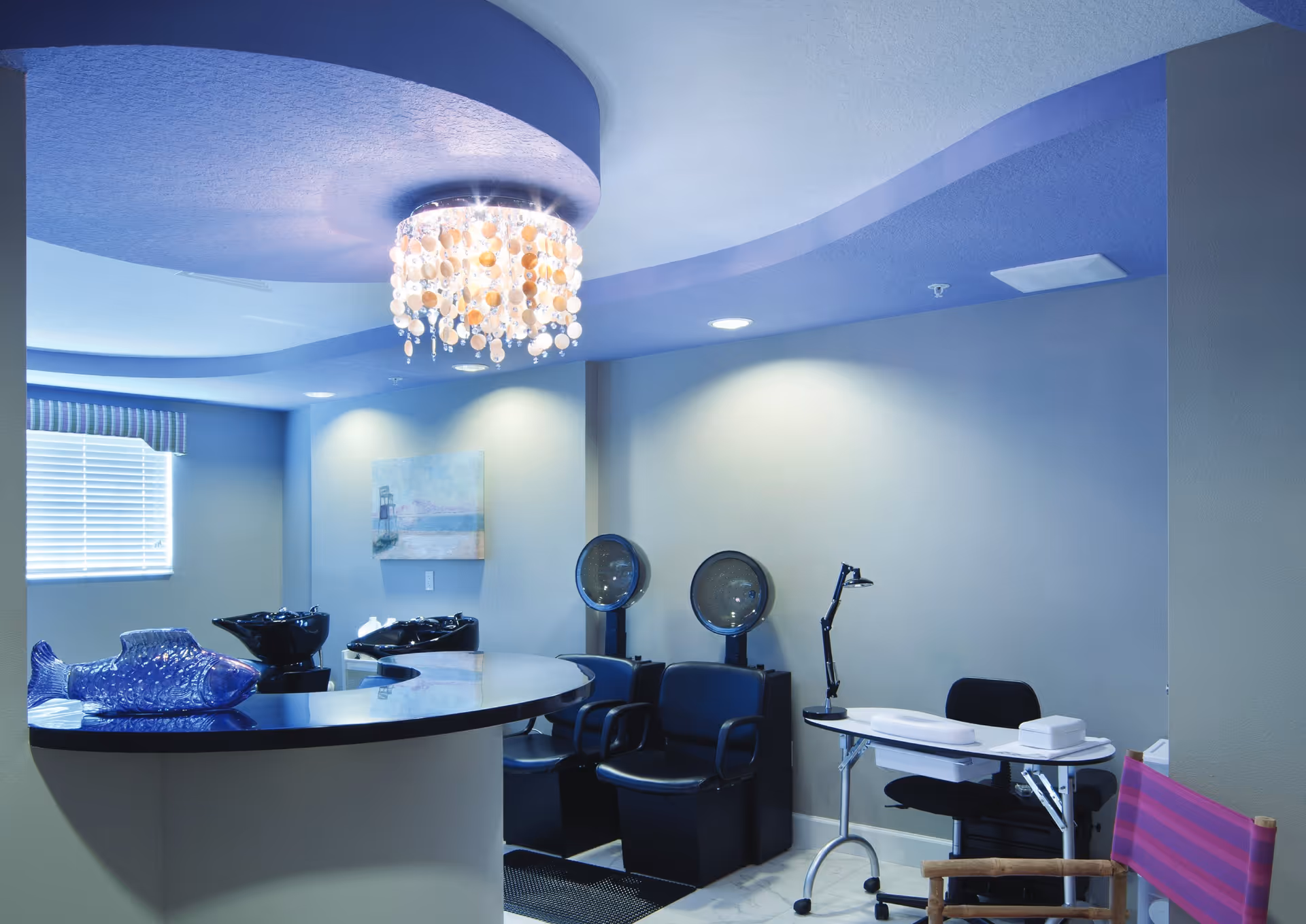 Interior salon room with hair-washing sinks, hooded dryers, styling chairs, a manicure table, and a decorative chandelier.