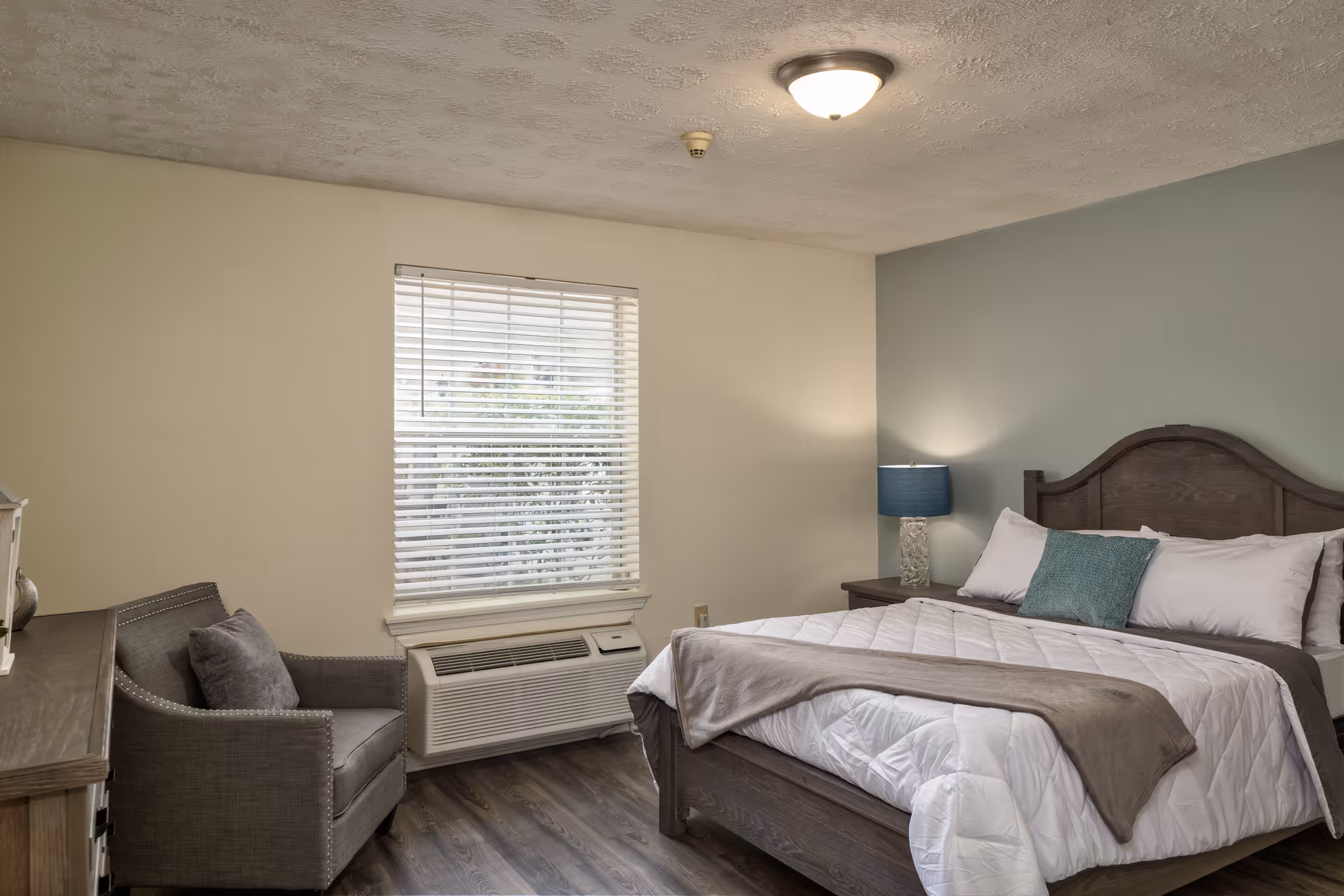 A cozy bedroom with a wooden bed frame, white bedding, a gray throw blanket, and a teal accent pillow. There is a wooden nightstand with a lamp featuring a blue shade. A gray armchair with a matching pillow sits next to a window with white blinds. The room has light-colored walls and wood flooring.